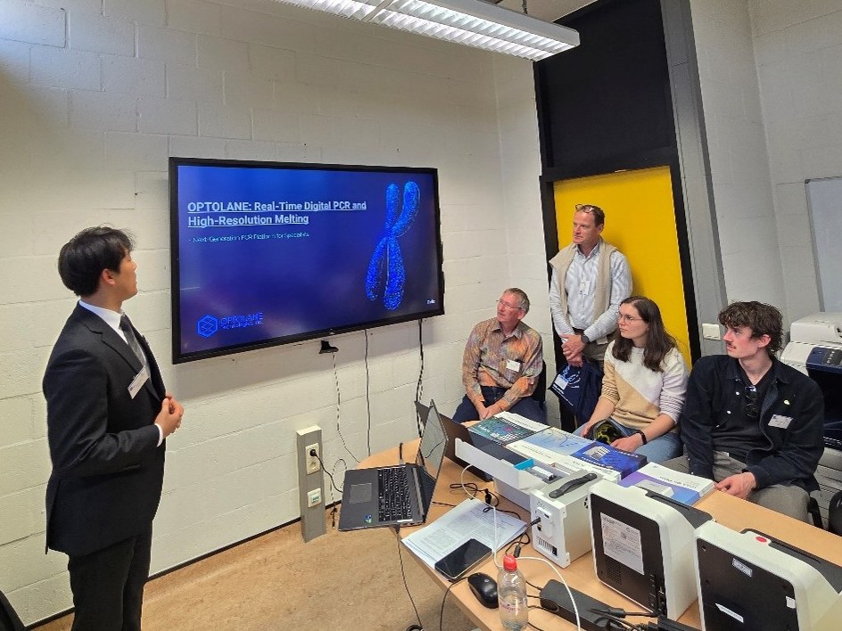 Jinwoo Seol presenting beside the OPTOLANE title slide to four listeners, with LOAA instruments on the table